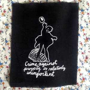 May include: Black and white embroidered patch featuring a silhouette of a person throwing a brick at a building. The text reads "Crime against property is relatively unimportant."