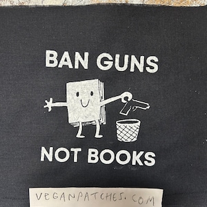 Könnte beinhalten: Schwarz-weißes Bügeleisen-Patch mit einer Comicfigur, die eine Waffe in einen Mülleimer wirft. Der Text lautet "Ban Guns Not Books" und "VeganPatches.com".