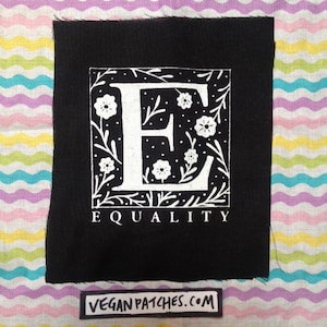以下が含まれることがあります： 白い花とツルで囲まれた白い文字Eが描かれた黒いアイロン接着パッチ。文字Eの下に「EQUALITY」と印刷されています。