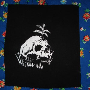 Op de afbeelding: Zwarte stoffen patch met een witte schedel en een klein plantje dat uit de schedel groeit.