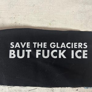 May include: Black fabric with white text that reads "SAVE THE GLACIERS BUT FUCK ICE". The fabric has a raw edge and is likely a patch or a piece of material for a garment or accessory.