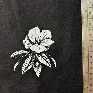 May include: A white flower with five petals and a yellow center, with leaves, printed on a black fabric background.