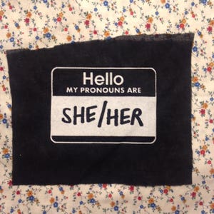 Op de afbeelding: Zwarte stoffen patch met een wit naamschild waarop staat "Hallo, mijn voornaamwoorden zijn Zij/Zij".