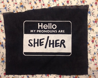 May include: Black fabric patch with a white name tag that reads "Hello, My Pronouns Are She/Her".