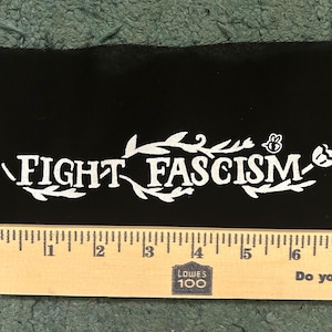 May include: A black fabric patch featuring the white printed text FIGHT FASCISM, surrounded by decorative leaf and flower illustrations and a small bee. A wooden ruler is positioned along the bottom edge, showing a length of approximately 8 inches.