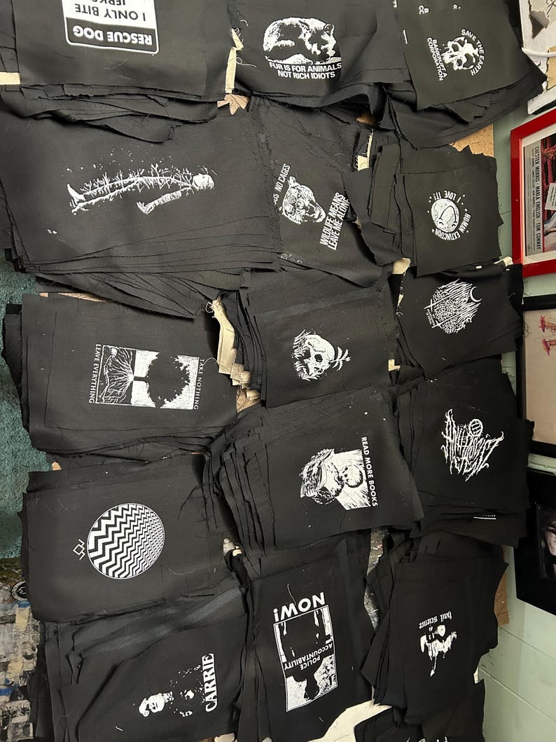 May include: A stack of black fabric patches with white text and images. The patches feature various slogans and designs, including "Rescue Dog", "Fur is for Animals, Not Rich Idiots", "Wildlife Means Leave Me Alone", "Read More Books", "Police Accountability", and "Carrie".