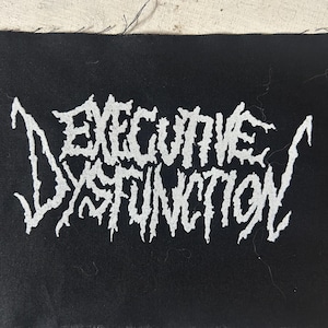 May include: Black rectangular patch with the words "EXECUTIVE DYSFUNCTION" in white, stylized, jagged lettering. The patch has a raw edge and is likely for sewing onto clothing or accessories.