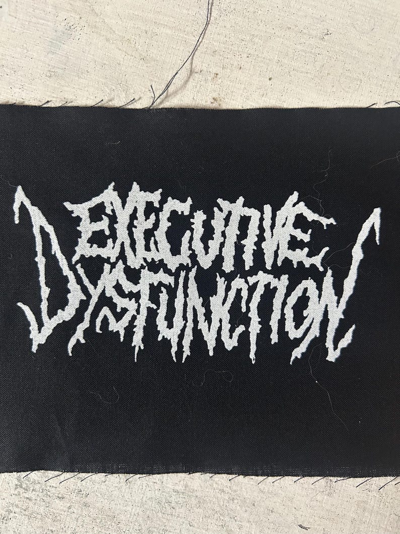 May include: Black rectangular patch with the words "EXECUTIVE DYSFUNCTION" in white, stylized, jagged lettering. The patch has a raw edge and is likely for sewing onto clothing or accessories.