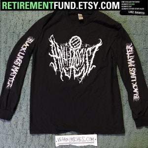 May include: Black long-sleeve shirt with a white band on the left sleeve that says "BLACK LIVES MATTER" and a white band on the right sleeve that says "BLACK LIVES MATTER". The shirt has a white graphic on the front that says "ANNIHILATION". The shirt is on a green surface. The text "RETIREMENTFUND.ETSY.COM" is at the top of the image. The text "VEGAN PATCHES.COM" is at the bottom of the image.