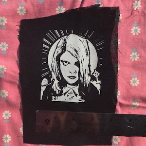 Puede incluir: Parche termoadhesivo en blanco y negro con una mujer de pelo largo, que lleva una camisa blanca con cuello. La mujer está rodeada por un halo y una cruz. El parche está sobre un fondo de tela negro.