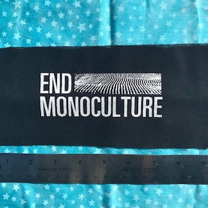 May include: Black fabric patch with white text that reads "END MONOCULTURE". The text is in a bold, sans-serif font. The patch is rectangular and has a rough, textured surface.