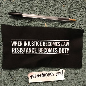 Puede incluir: Parche de tela negro con texto blanco que dice "When injustice becomes law resistance becomes duty". El parche está sobre una superficie verde y peluda. Un pequeño trozo de papel blanco con escritura negra que dice "veganpatches.com!" está en la esquina inferior derecha de la imagen.