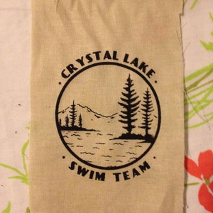 以下が含まれることがあります： ベージュの布のパッチで、黒いインクで「クリスタルレイクスイムチーム」という文字が円形に書かれています。円の内部には、山と3本の松の木のある湖の黒いシルエットがあります。