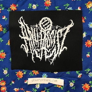 Peut inclure: Un écusson noir avec un texte blanc stylisé et un motif circulaire en haut. Le texte semble être le nom d'un groupe ou un logo. L'écusson est posé sur un tissu bleu à motifs floraux. Une petite étiquette blanche en bas indique "veganpatches.com".