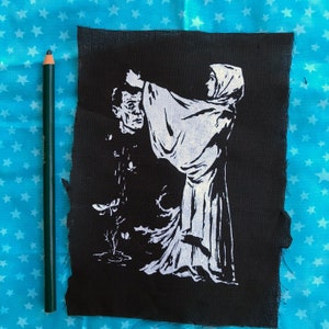 May include: Black fabric patch with a white graphic depicting a figure in a hooded robe holding a severed head. A green pencil is next to the patch. The background is a blue fabric with white stars.