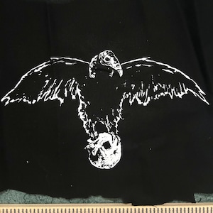 May include: A black fabric patch featuring a white screen-printed graphic of a vulture with wings spread wide, clutching a human skull in its talons. A wooden ruler is positioned at the bottom, showing a width of approximately 15 cm.