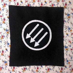 Puede incluir: Parche de tela negro con un círculo blanco que contiene tres flechas blancas que apuntan hacia la izquierda. Las flechas están dispuestas en línea vertical.