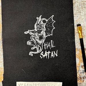 May include: Black iron-on patch with a white image of a winged demon and the text "Hail Satan". The patch is on a black fabric background. The text "Vegan Patches, CRM" is visible on a white label below the patch.