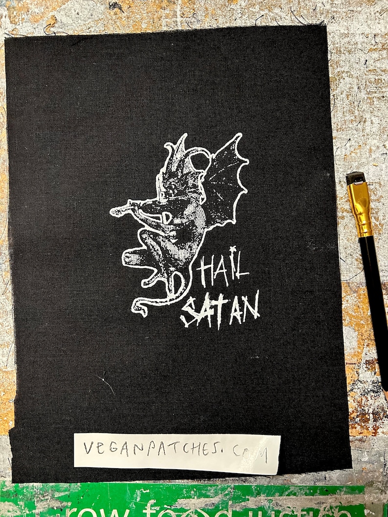 May include: Black iron-on patch featuring a white image of a winged demon and the text "Hail Satan". The patch is on a black fabric background. The text "Vegan Patches, CRM" is visible on a white label below the patch.