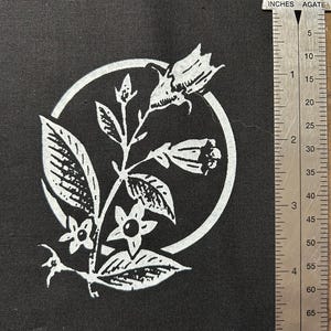 May include: Black fabric square with a white floral design inside a circle. The design includes leaves, flowers, and buds. A metal ruler with inch measurements is on the right side of the fabric.