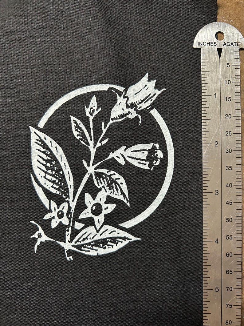May include: Black fabric square with a white floral design inside a circle. The design includes leaves, flowers, and buds. A metal ruler with inch measurements is on the right side of the fabric.