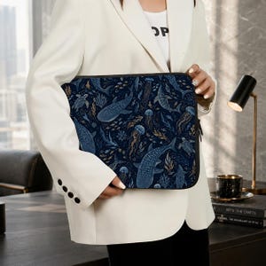 May include: A dark blue laptop sleeve featuring a marine life pattern. The design includes whale sharks, jellyfish, and seaweed in shades of blue and gold. The sleeve has a black zipper and is held by a person wearing a white blazer.