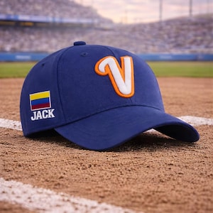 Op de afbeelding: Een marineblauwe baseballpet met een gebogen klep. De pet heeft een grote witte en oranje letter "V" op de voorkant en een kleine vlag en de naam "JACK" aan de zijkant. De pet ligt op een honkbalveld.