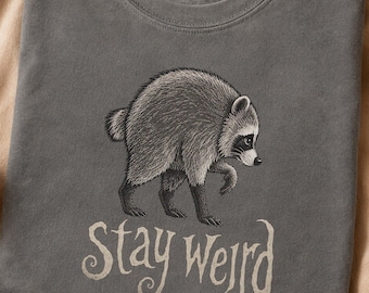 May include: A charcoal gray crew neck t-shirt featuring a detailed black and white illustration of a raccoon walking. Below the raccoon, the text "Stay Weird" is printed in a stylized, flowing white font. The shirt has a soft, washed-out texture.