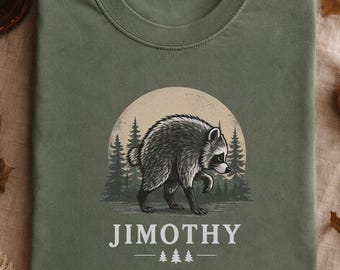 May include: An olive green t-shirt featuring a graphic of a raccoon standing in front of a large moon and a forest of pine trees. Below the raccoon, the text JIMOTHY is printed in white capital letters, flanked by two small pine tree icons.