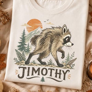 Pode incluir: Uma t-shirt de cor creme com um gráfico de um guaxinim a caminhar por uma floresta com montanhas e um sol ao fundo. O texto "JIMOTHY" está impresso numa fonte castanha escura por baixo do guaxinim, acompanhado por um pequeno ícone de pinheiro.