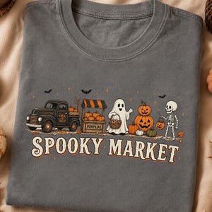 Pode incluir: Uma t-shirt cinzento-carvão com um gráfico de uma carrinha preta, uma banca de abóboras, um fantasma, uma pilha de abóboras e um esqueleto. O texto SPOOKY MARKET está impresso em cor creme sob a ilustração com pequenos morcegos.