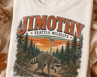 May include: A cream-colored t-shirt featuring a graphic print with the text "JIMOTHY" and "SEATTLE WILDLIFE" above a forest scene. The illustration depicts a raccoon walking on a path with pine trees, a sunset, and the Space Needle.