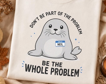 May include: A cream-colored t-shirt featuring a graphic of a gray seal wearing a name tag that reads "HELLO NEIL". The text above the seal says "DON'T BE PART OF THE PROBLEM" and the text below the seal says "BE THE WHOLE PROBLEM" in black.