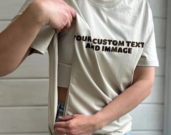 May include: A beige t-shirt with the text "YOUR CUSTOM TEXT AND IMMAGE" in brown. The shirt has a hidden pocket. The person is wearing blue jeans.