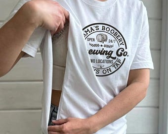 May include: White t-shirt with a round neck featuring a black circular logo that reads "Mama's Boobery Brewing Co." with a barrel illustration. The shirt is being lifted to reveal a white top and blue jeans.
