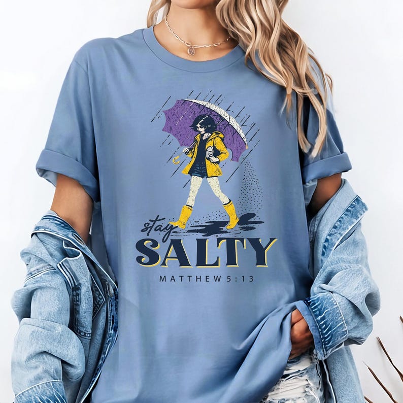 May include: A light blue t-shirt with the words "stay SALTY" and "MATTHEW 5:13" printed on the front. The graphic features a person in a yellow raincoat and boots holding a purple umbrella in the rain.