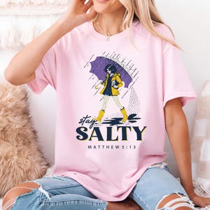 May include: Pink t-shirt with a graphic of a person walking in the rain under an umbrella. The person is wearing a yellow raincoat and boots. The text "stay SALTY" and "MATTHEW 5:13" are printed below the graphic.