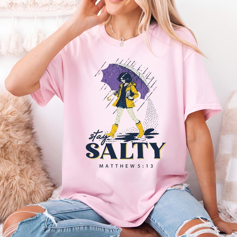 May include: Pink t-shirt with a graphic of a person walking in the rain under an umbrella. The person is wearing a yellow raincoat and boots. The text "stay SALTY" and "MATTHEW 5:13" are printed below the graphic.