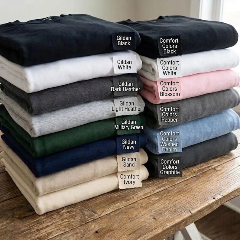 May include: A stack of folded sweatshirts in various colors, including black, white, gray, and navy. Each sweatshirt has a tag indicating the color name, such as "Gildan Black" and "Comfort Colors White". The sweatshirts are neatly arranged on a wooden surface.
