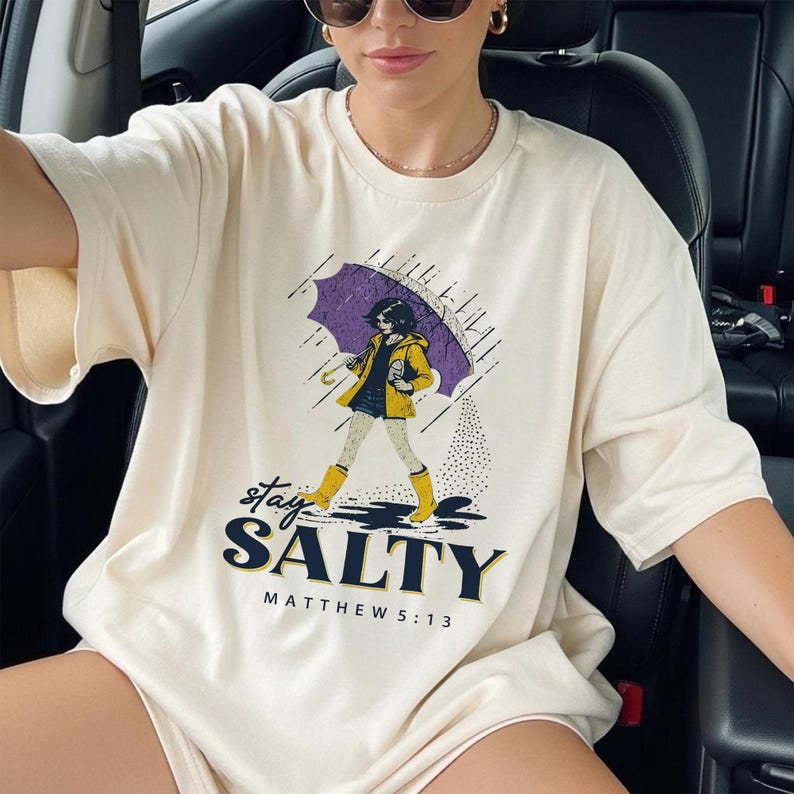 May include: Cream-colored t-shirt featuring a graphic of a person under a purple umbrella in the rain. The text "stay SALTY" and "MATTHEW 5:13" are printed below the graphic. The person is wearing a yellow jacket and boots.