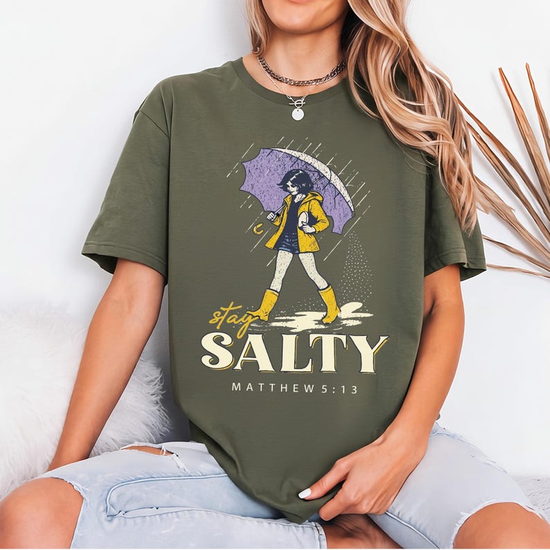 May include: Olive green t-shirt featuring a graphic of a person walking in the rain under a purple umbrella, wearing a yellow jacket and boots. The text "stay SALTY" and "MATTHEW 5:13" are below the graphic.