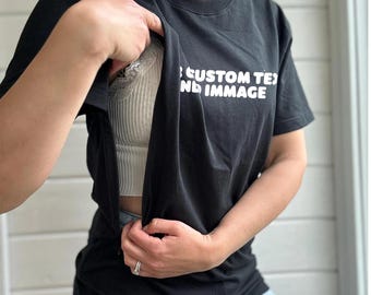 May include: Black crew neck t-shirt with white text that reads "CUSTOM TEXT AND IMMAGE". The shirt is being held open to reveal a white ribbed tank top. The model is wearing blue jeans.