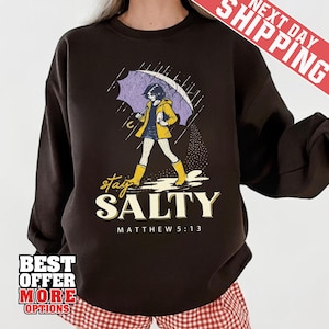 May include: Dark brown sweatshirt with a graphic of a person walking in the rain under an umbrella. The person is wearing a yellow jacket and boots. The text "stay SALTY" and "MATTHEW 5:13" are below the graphic.