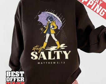 May include: Dark brown sweatshirt with a graphic of a person walking in the rain under an umbrella. The person is wearing a yellow jacket and boots. The text "stay SALTY" and "MATTHEW 5:13" are below the graphic.