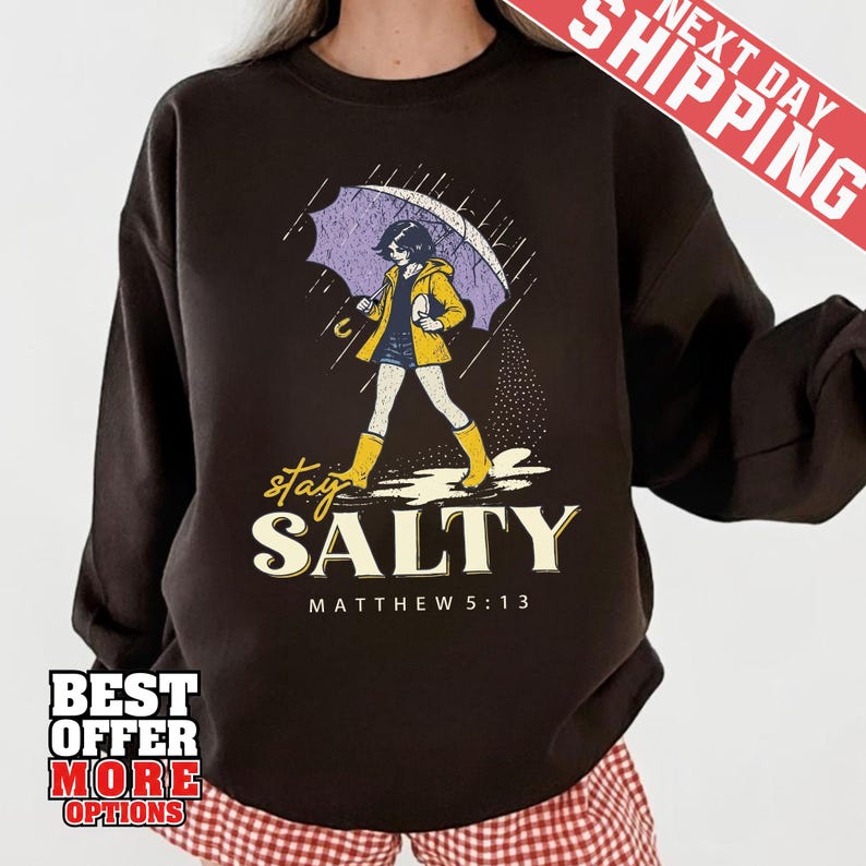 May include: Dark brown sweatshirt with a graphic of a person walking in the rain under an umbrella. The person is wearing a yellow jacket and boots. The text "stay SALTY" and "MATTHEW 5:13" are below the graphic.