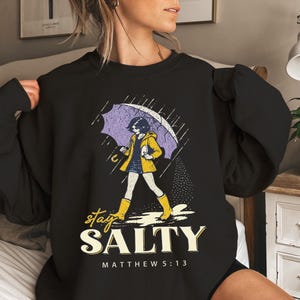 May include: Black sweatshirt with a graphic of a person walking in the rain under an umbrella. The person is wearing a yellow jacket and boots. The text "stay SALTY" and "MATTHEW 5:13" are below the graphic.