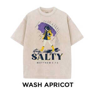 May include: Apricot-colored t-shirt with a graphic of a person walking in the rain under a purple umbrella. The graphic includes the words "stay SALTY" and "MATTHEW 5:13". The shirt is a crew neck style.