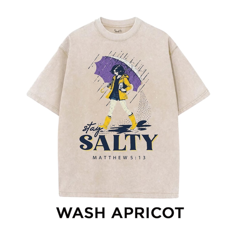 May include: Apricot-colored t-shirt with a graphic of a person walking in the rain under a purple umbrella. The graphic includes the words "stay SALTY" and "MATTHEW 5:13". The shirt is a crew neck style.