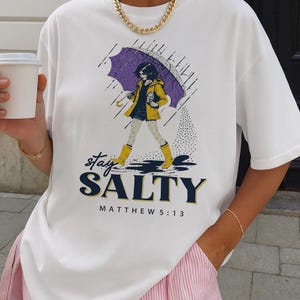May include: White t-shirt featuring a graphic of a person walking in the rain under an umbrella. The person is wearing a yellow jacket and boots. The text "Stay Salty" and "Matthew 5:13" are printed on the shirt. A gold chain necklace is also visible.