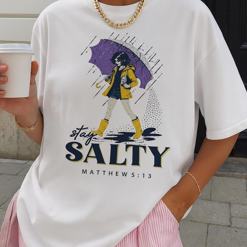 May include: White t-shirt featuring a graphic of a person walking in the rain under an umbrella. The person is wearing a yellow jacket and boots. The text "Stay Salty" and "Matthew 5:13" are printed on the shirt. A gold chain necklace is also visible.